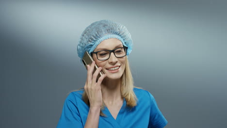 Caucasian-attractive-young-female-medic-in-blue-costume,-hat-and-glasses-speaking-cheerfully-on-the-phone-and-smiling.